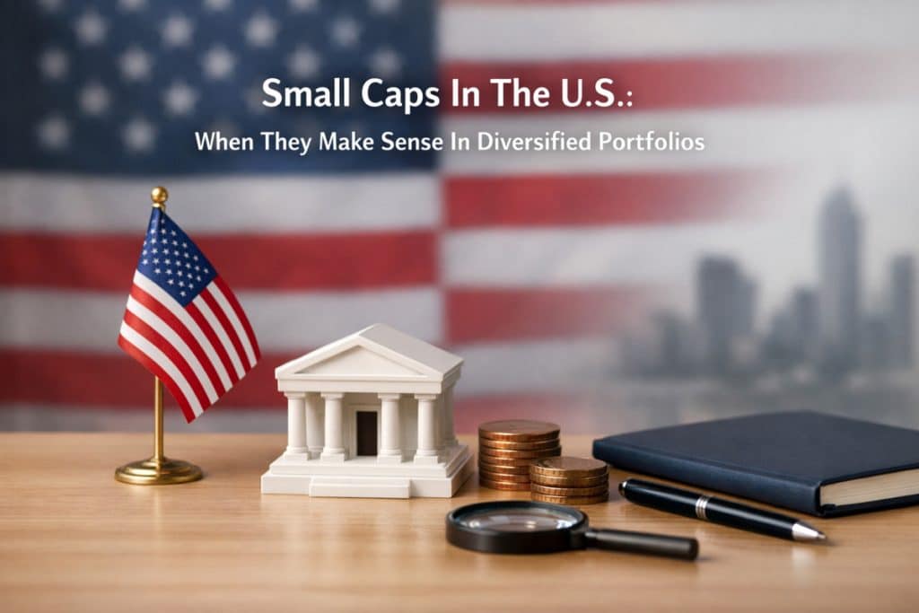Cover image titled ‘Small Caps In The U.S.: When They Make Sense In Diversified Portfolios’ over a blurred U.S. flag background and a faint city skyline. On a wooden desk in the foreground are a small U.S. flag on a stand, a white classical building model, stacks of coins, a magnifying glass, a closed notebook, and a pen—suggesting research and evaluation of small-cap investments in a diversified portfolio.